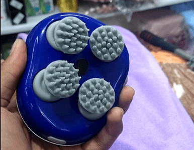 Hand holding a blue handheld electric massager with four rotating silicone massage heads displayed in a store